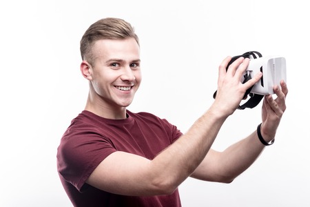 Excited gamer. Upbeat fair-haired young man in a burgundy t-shirt being about to put on a VR headset and smiling at the camera happily while posing against a white backgroundの写真素材