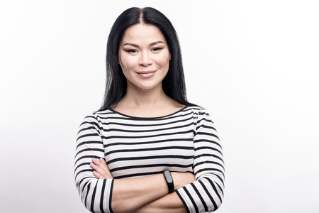 Charming female. Attractive young woman in a striped pullover posing for the camera while folding her arms across her chest, having a fitness tracker on her wristの写真素材