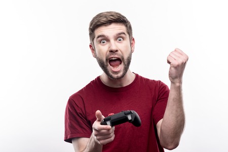 Enthusiastic gamer. Joyful bristled young man holding a video game controller and raising his hand triumphantly, celebrating victory in game, while posing isolated on a white backgroundの写真素材
