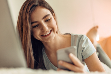Texts full of love. The close up of a charming teenage girl checking the notifications on her phone, reading the messages and smiling happily while lying on the bedの写真素材