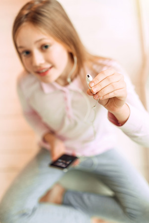 Listen with me. The top view of a charming teenage girl sitting cross-legged and smiling at the camera while giving an earphone, being in the focusの写真素材