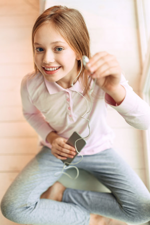 Give it a try. Top view of a cute teenage girl sitting cross-legged on the floor and suggesting listening to music, handing one earphoneの写真素材