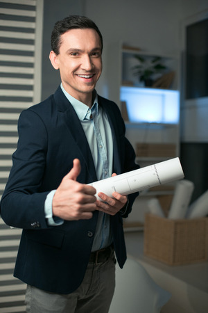 Excellent project. Enthusiastic joyful male architect showing thumb while smiling to the camera and carrying blueprintの写真素材