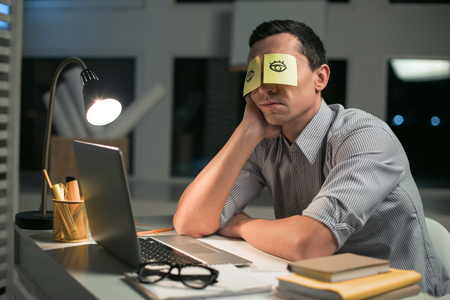 Office fun. Exhausted creative male employee placing stickers while touching his face and sitting at the tableの写真素材