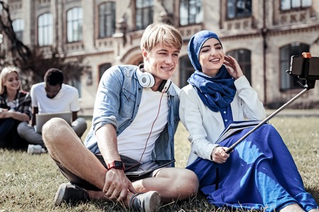No prejudices. Positive nice religious woman holding a selfie stick and taking a photo while sitting on the grass with her friendの写真素材