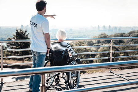 How beautiful. Nice positive handsome man looking at the view and pointing with his hand while having a walk with a disabled elderly womanの写真素材