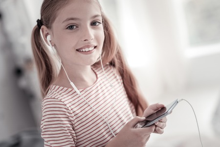 Music lover. Cheerful lovely long-haired girl with pig-tails smiling and wearing a headset and holding her phone while listening to the musicの写真素材