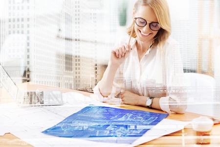 Bright idea. Young smiling designer wearing glasses and looking at the sketch while sitting at the tableの写真素材