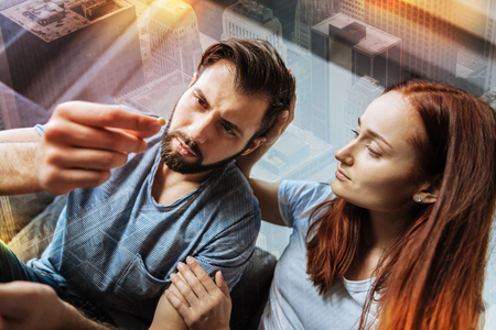 Medical help. Tired young unhappy man feeling awful while looking at the pill and his girlfriend sitting near and supporting himの写真素材