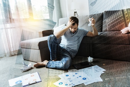 Furious. Young emotional angry man feeling terrible and scrunching paper while sitting on the floor...