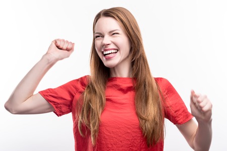 Emotional reaction. Upbeat ginger-haired young woman raising her hands in a gesture of celebration and laughing happily, having heard good newsの写真素材
