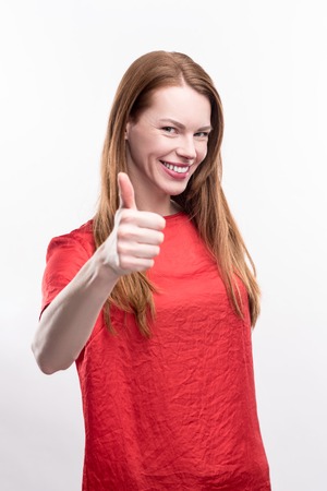 Sign of approval. Pretty ginger-haired young woman in a red t-shirt showing a thumbs up and smiling at the camera while posing against a white backgroundの写真素材
