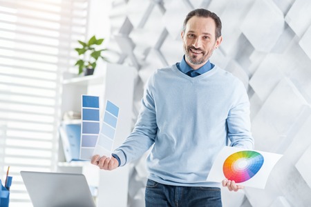 Happiness. Attractive happy dark-haired bearded man smiling and holding some sheets of paper while standing in his officeの写真素材