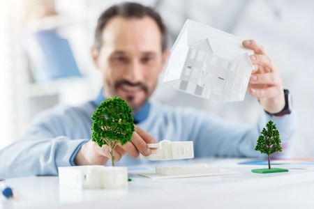 I love constructing. Good-looking cheerful dark-haired bearded man smiling and holding a model of a future house while sitting at the tableの写真素材