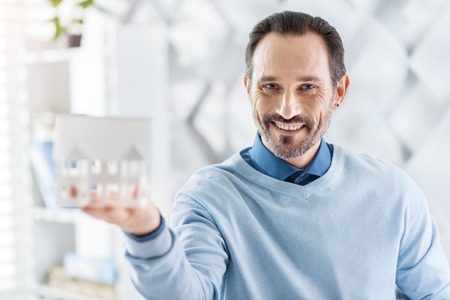 Successful project. Handsome alert dark-haired bearded man holding the model of the house and smiling and showing itの写真素材