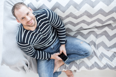 Being energetic. Good-looking cheerful bearded young man smiling and sitting on the couch and holding his phoneの写真素材