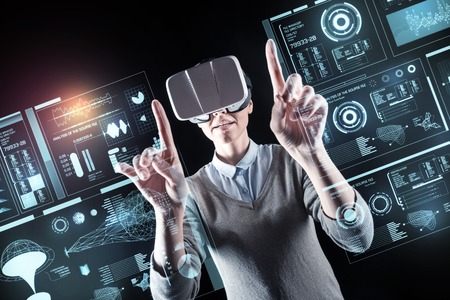 Clever young experienced programmer feeling curious while wearing virtual reality glasses and touching the screen of a futuristic deviceの写真素材