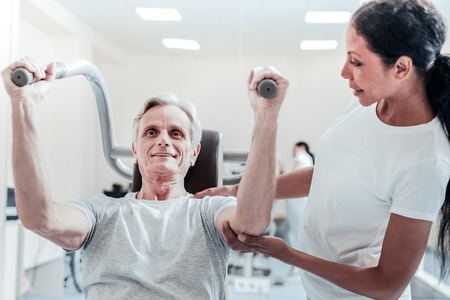 Exuberant energetic old grey-haired man exercising on a training device and a pretty young dark-haired female trainer standing near him and touching his handの写真素材