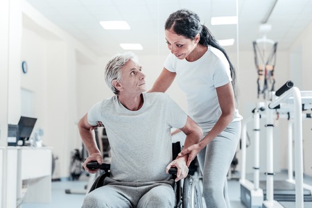 Concentrated old grey-haired man sitting in a wheelchair and an attractive young dark-haired woman standing near him and smiling while they looking at each otherの写真素材