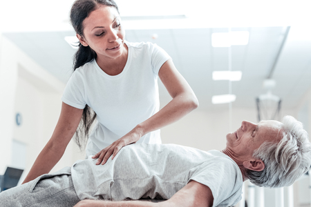 Vigorous old wrinkled grey-haired man lying on a ball for exercises and a joyful young dark-haired afro-american woman helping him and touching his stomachの写真素材