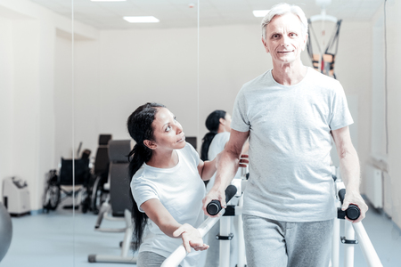 Vigorous old wrinkled grey-haired man smiling and exercising on a training device while a young dark-haired female trainer smiling and looking at himの写真素材