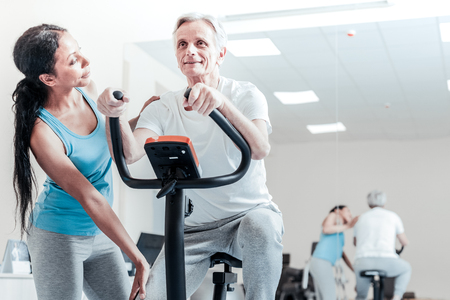Alert smiling aged grey-haired man exercising on a training device while a beautiful serious young dark-haired afro-american female trainer standing near him and looking at himの写真素材