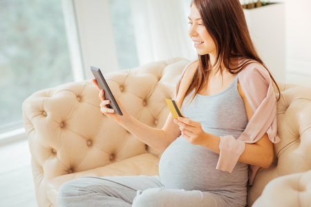 Joyful parturient smart woman sitting on the sofa using the tablet and holding credit card.の写真素材