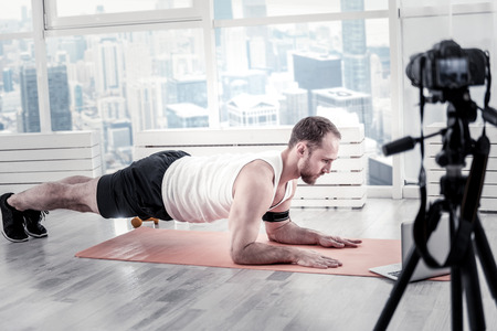 Attractive bearded male blogger staying in plank while posing near window and using cameraの写真素材