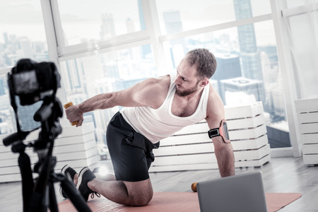 Concentrated focused male blogger using dumbbell while looking at it and exercising in front of...
