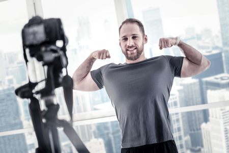 Vigorous charming male blogger showing biceps while sharing his fitness program and using cameraの写真素材