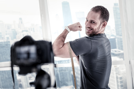 Vigorous joyful male blogger measuring biceps size while grinning and updating blogの写真素材