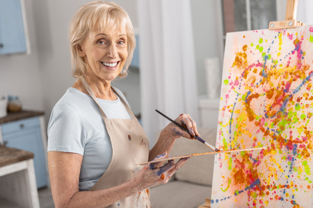 Joyful jolly mature woman carrying palette and tassel while grinning and staring at cameraの写真素材