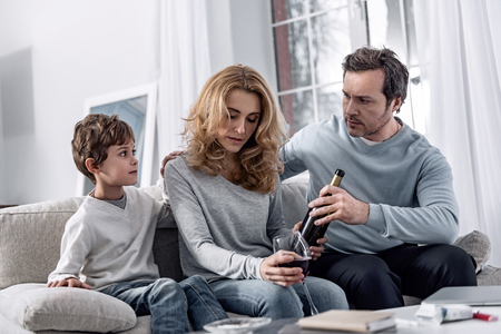 Young beautiful serious woman with a bottle of wine in her hand understanding her alcohol addiction and feeling guilty while her family members sitting nearの写真素材