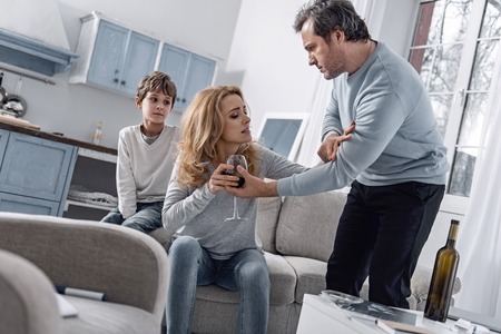 Emotional serious worried man looking at his alcohol addicted wife and insisting on consulting a doctor while trying to take a glass of wine from her hand with their son watchingの写真素材