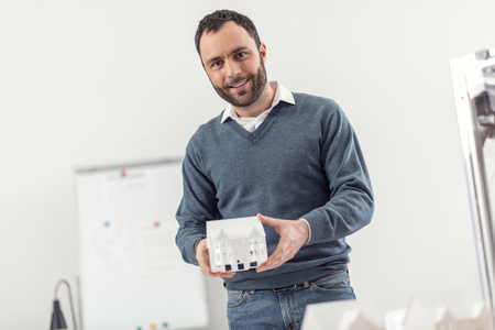 Happy engineer. Upbeat young man working as an engineer posing for the camera while showing a miniature model of a house, having printed it with a 3D printerの写真素材
