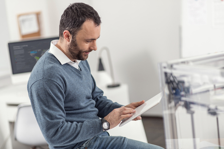 Busy worker. Pleasant bearded young man sitting on his work desk near the 3D printer and working on his tablet, being focused on the taskの写真素材