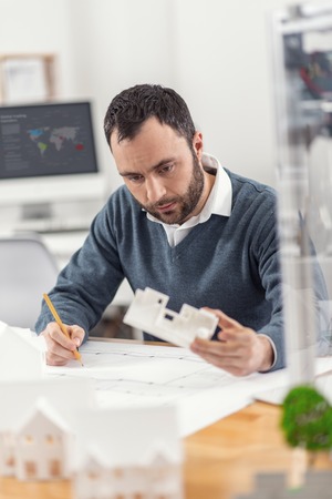 Important changes. Handsome dark-haired young engineer holding a 3D model and making corrections in the blueprint according to its construction, being focused on the taskの写真素材