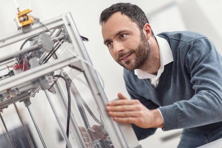 Careful treatment. Charming bearded young man touching the body of a 3D printer and trying to move...
