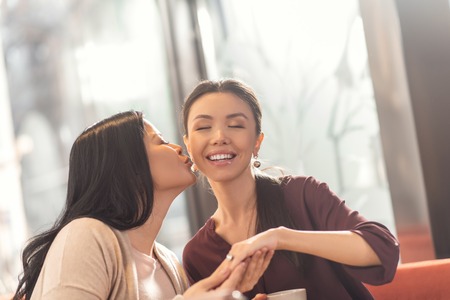Be happy. Positive nice joyful woman sitting together with her friend and kissing her while being happy about her engagementの写真素材