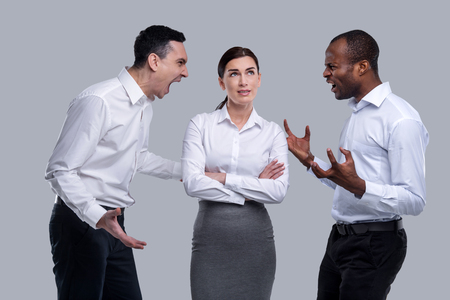 Teamwork. Pretty thoughtful dark-haired young woman thinking and standing between two angry men and...