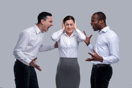 Quarrelling. Pretty annoyed dark-haired young woman closing her ears with her hands and standing between two angry men and men shouting at each otherの写真素材