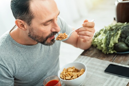Occupied concentrated calm man sitting in the kitchen by the table opening mouth and eating.の写真素材