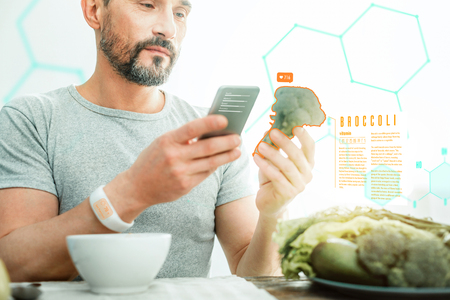 Handsome interested occupied man standing with the cellphone in his hand holding and examining a broccoli.の写真素材