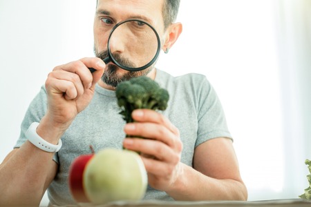 Curious searching concentrated man spending time in the bright room holding a broccoli looking through magnifying glass.の写真素材