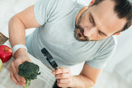 Pleasant curious man being in the bright room holding a broccoli using the magnifying glass.の写真素材