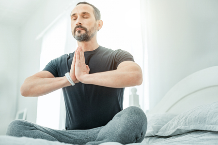 Calm healthy occupied man sitting in the bright room on the bed holding hands opposite his body closing his eyes.の写真素材