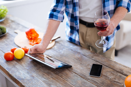 Close up of sophisticated tablet rising by male hands which holding glass of red tasty wine and phone lying on surfaceの写真素材