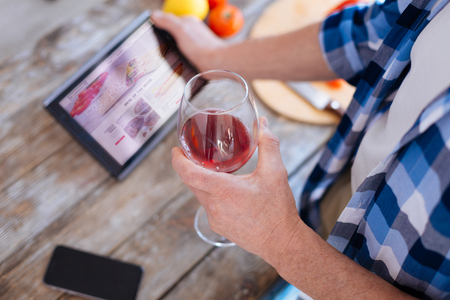 Selective focus of glass of exclusive red wine hold and elevated by male hand while tablet placed on surfaceの写真素材