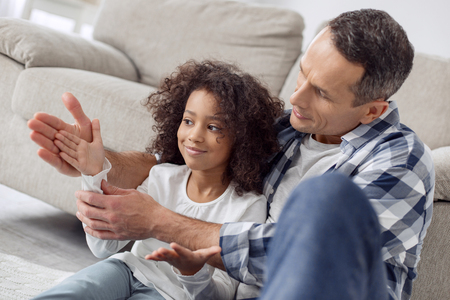Pretty cheerful curly-haired girl smiling and sitting with her father and he holding her hand in hisの写真素材