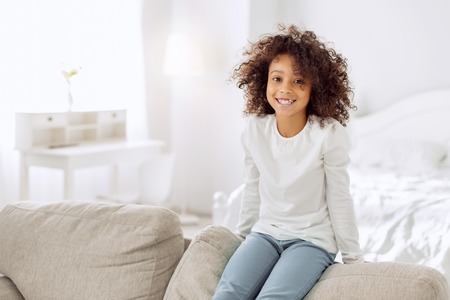 Pretty content curly-haired girl smiling and sitting on the couch and wearing a white shirt and jeansの写真素材